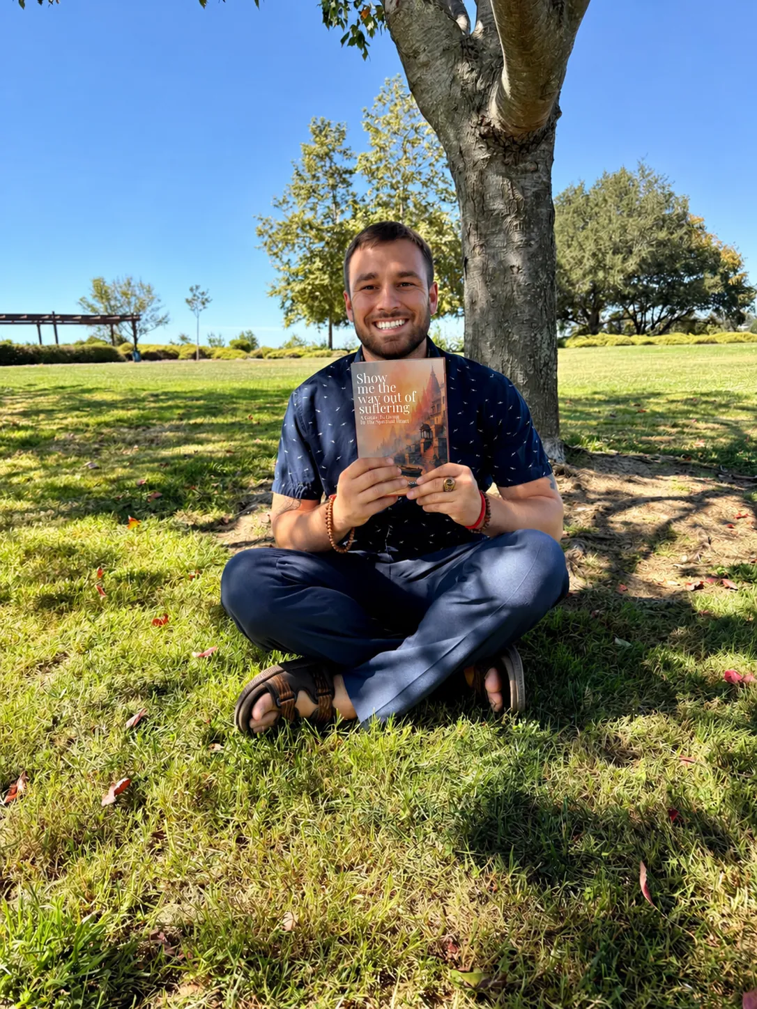 Alex Momcilovic sitting cross-legged under a tree, holding his book