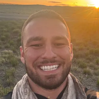 Alex Momcilovic smiling, with the sun setting over open country behind him